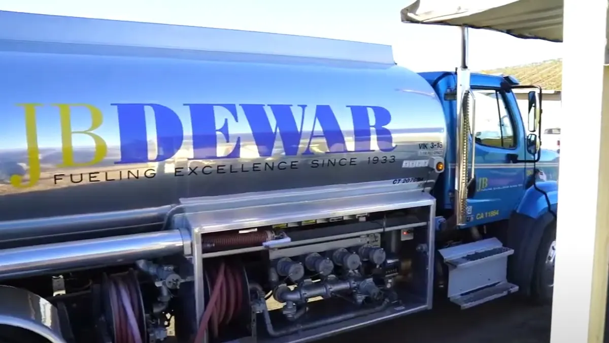 A screenshot of a video, showing a fuel tanker driving past a building. Text on the tanker says "JB Dewar Fueling Excellence since 1933."