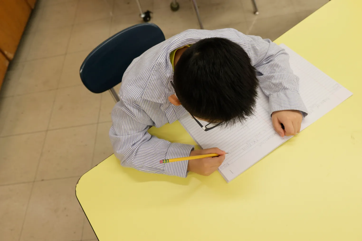 Student sits at desk with paper and pencil, looking down at the paper. Camera shot is from above student's head.