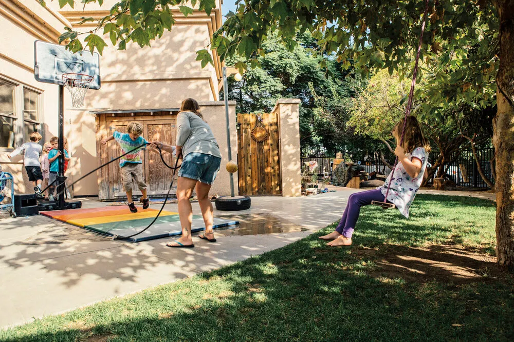 Outside in a yard, a caregiver swings a jump rope while a child jumps in the middle, while another child swings on a tree swing.