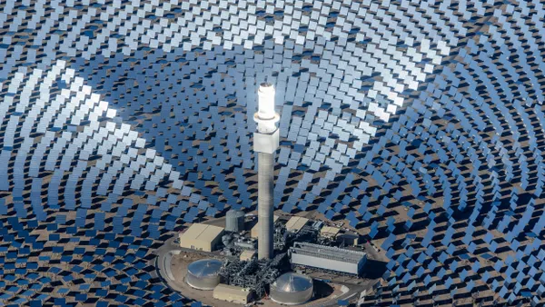 An aerial view of a solar power facility with a central tall white tower surrounded by concentric circles of blue reflective solar panels, set in a desert landscape.