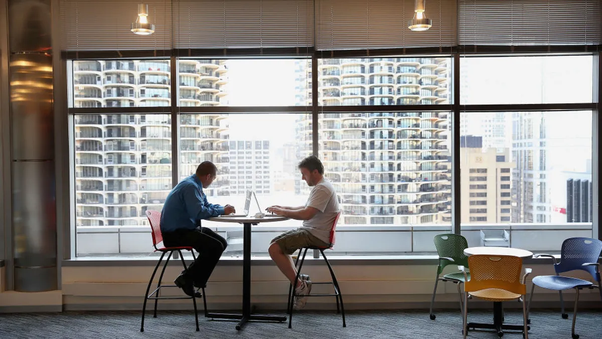 Two people on laptops sit across from each other at a table in front of a large window with Chicago skyscrapers in the background.