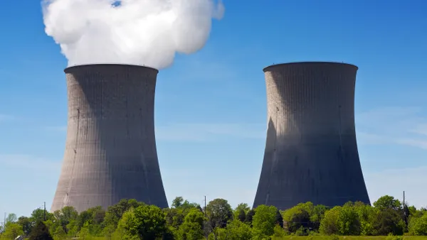 Two large hyperboloid cooling towers rise above a green tree-covered landscape. One tower emits a thick plume of white steam into a clear blue sky, while the other stands still. The towers are cylindrical at the base and narrow in the middle with a wider top.