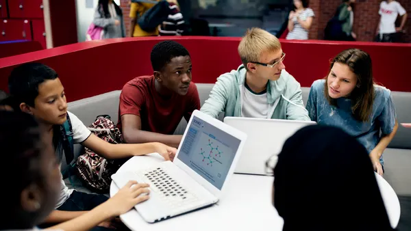 Six students are seated around a round table. Two people have open laptops on the table