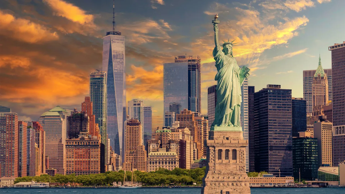 Statue of Liberty and New York City Skyline with Manhattan Financial District, Battery Park, Water of New York Harbor, World Trade Center, Sail Boat and Dramatic Orange and Blue Sunset Sky with Clouds