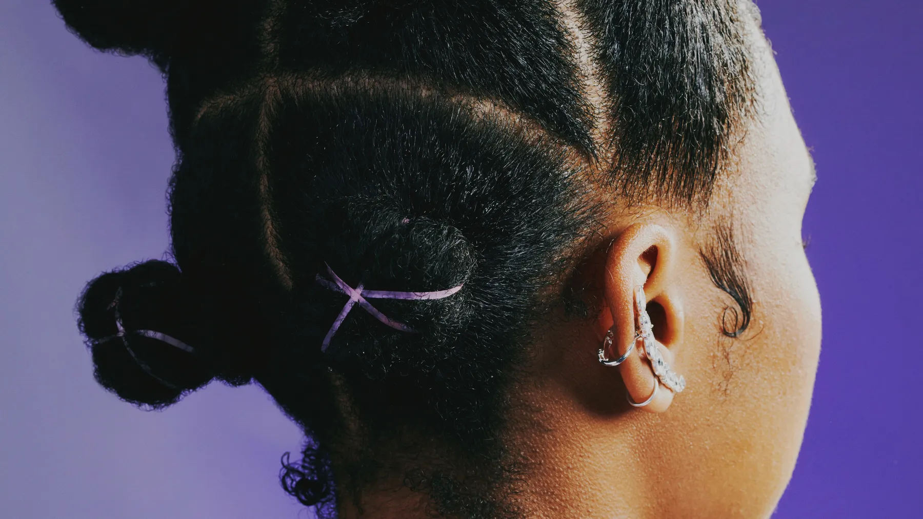 Close up: Bantu knots with purple elastics. A Black woman stands against a purple studio background, wearing a lighter purple windbreak that is zipped up on the back her neck