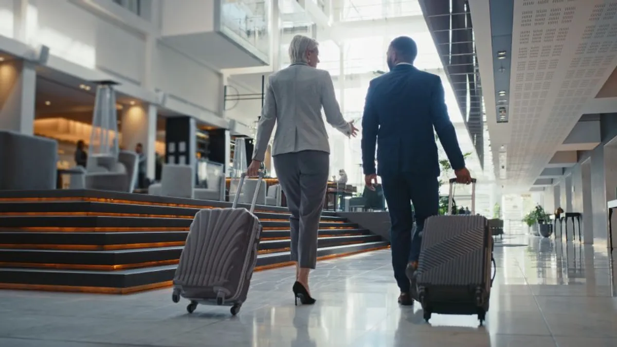 Business travelers walk through hotel lobby carrying luggage.