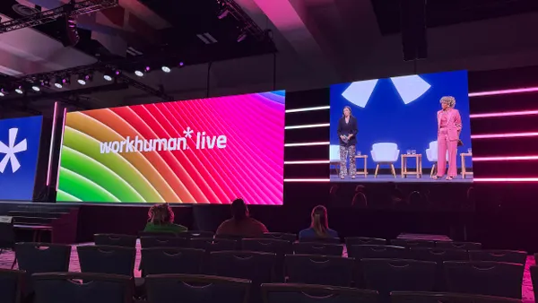 "Workhuman" is on a colorful background on a screen above a stage. To the right, two women present.