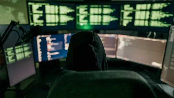 A person wearing a hoodie sits in front of multiple compluter screens with green code displayed.