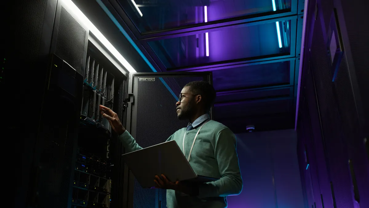 A data engineer working with a supercomputer in server room lit by blue light and holding laptop, copy space.