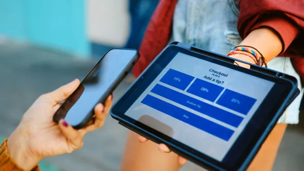 A woman holds up her smart phone to a tablet that is asking what percentage of a tip should be added.