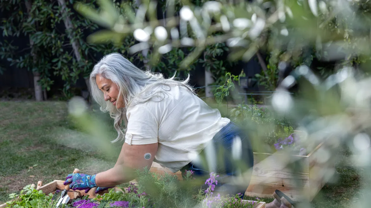 A person digs with a spade in a raised garden bed. They wear a gray sticker on their upper arm.