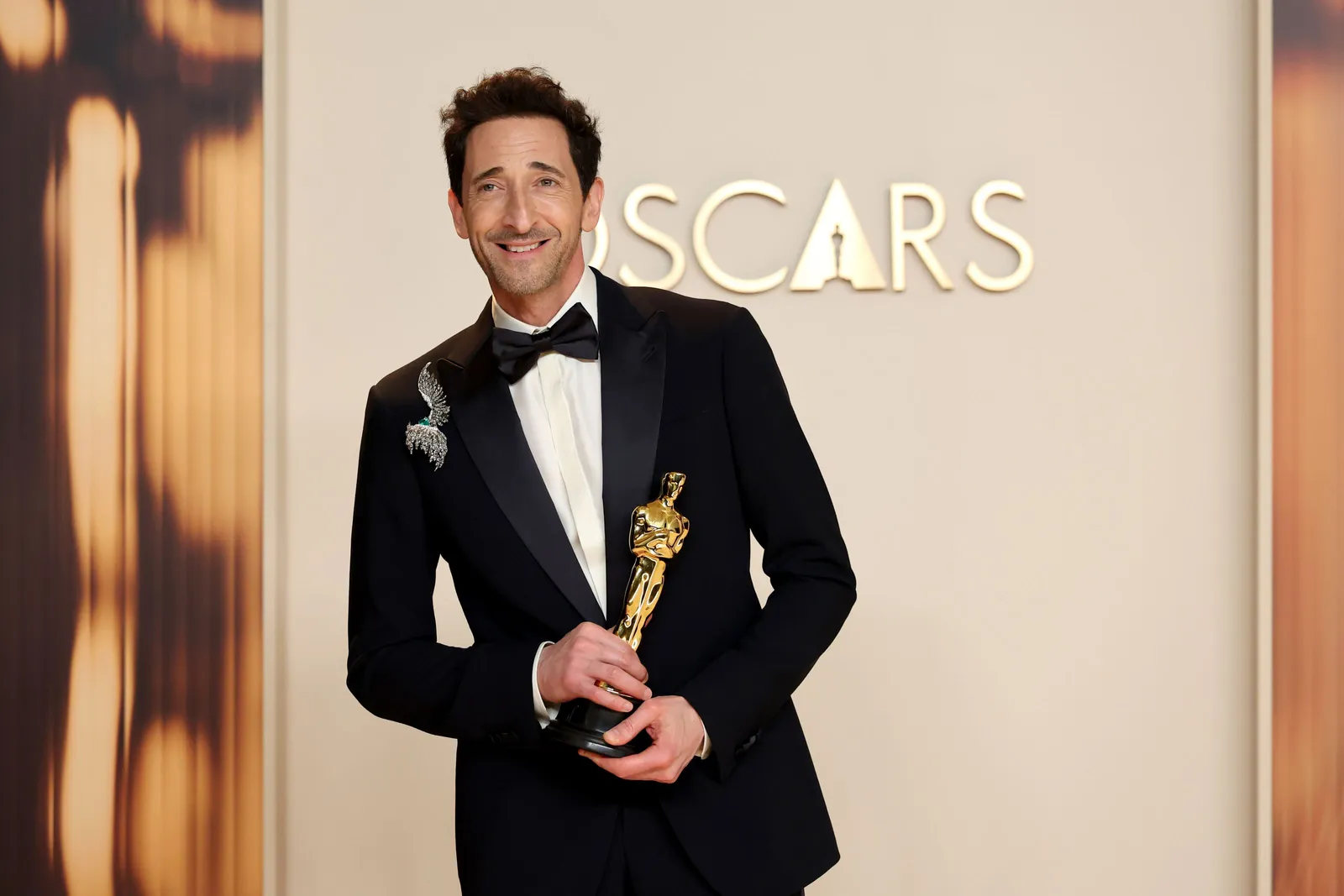 A man in a classic tuxedo embellished with a large diamond lapel pin holds an Oscar and stands on the red carpet.