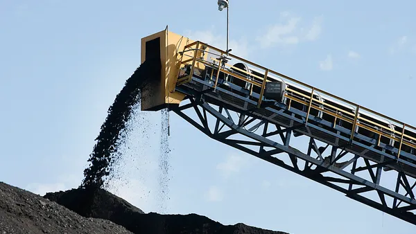 Coal falls off a conveyor belt onto a larger pile of coal. Multiple mounds of coal can be seen.