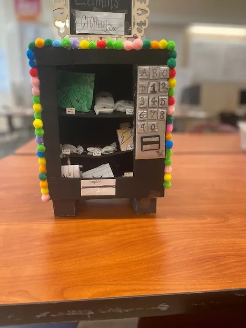 A miniature clothing vending machine sits on top of a desk in a classroom.