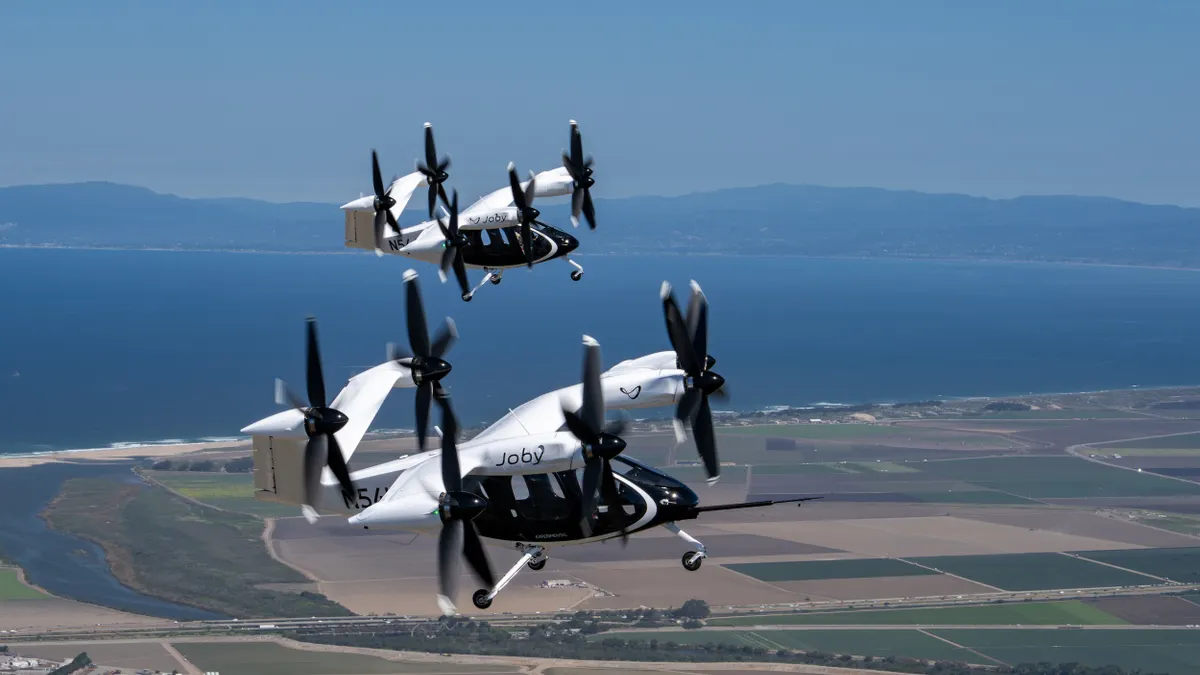 Two futuristic electric vertical takeoff and landing aircraft fly over farmland.