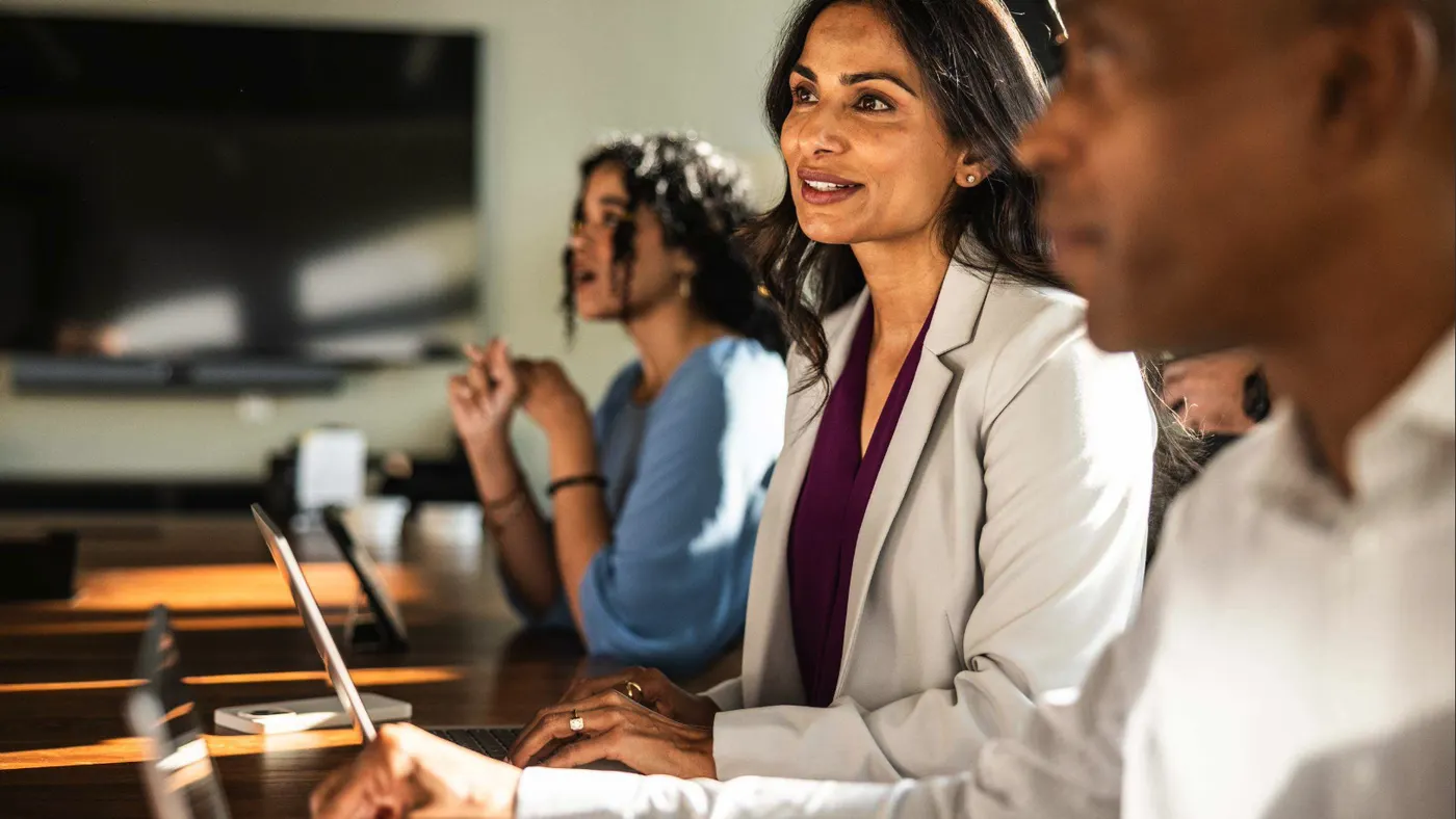 Three colleagues at a conference table with laptops, including a woman in a white blazer looking ahead and smiling.