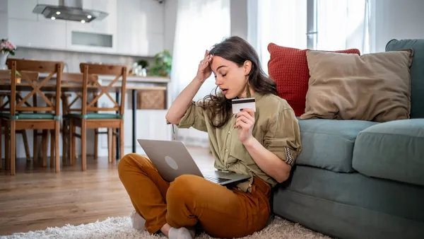 A young woman sits on the floor with a laptop on her legs and a credit card in her hand.