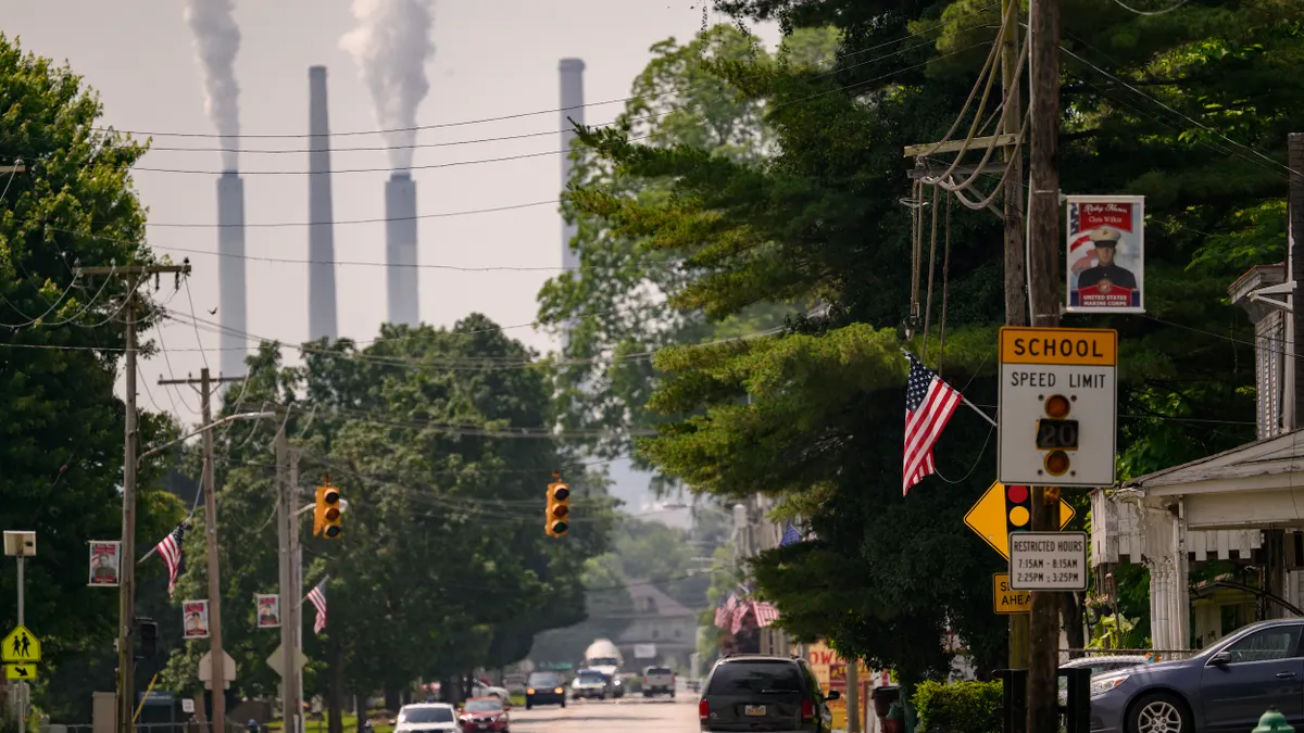 Cars on a city street with "School Speed Limit" sign and American flag in the foreground and four smokestacks in the background.
