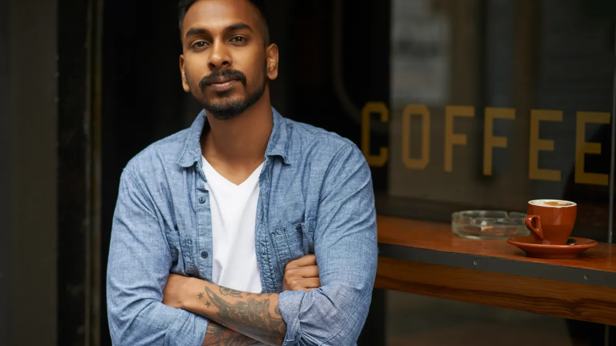 Portrait of a young South Asian man with tattoos, a beard and a mustache sitting outside a cafe