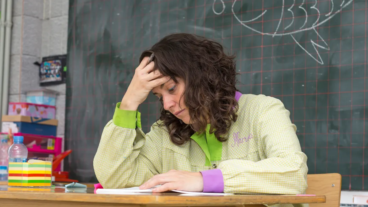 A person is sitting at a desk in a classroom. They look stressed. A chalkboard is behind them and there are papers on the desk.