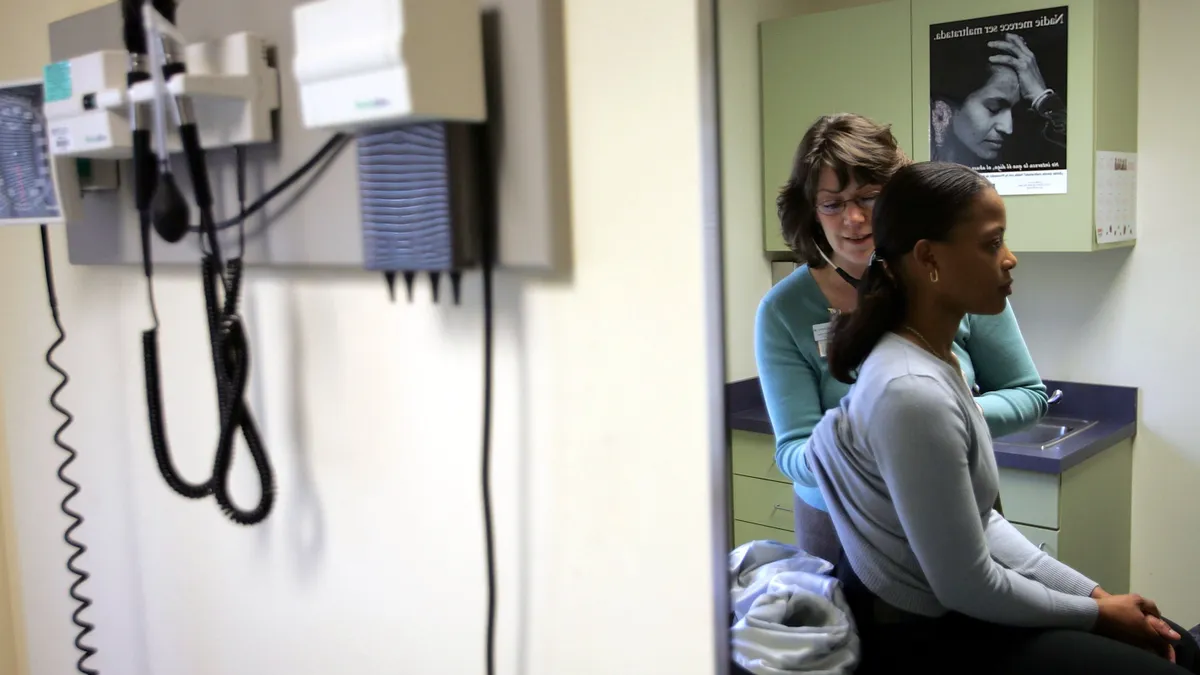 A patient sits on an examination table in a doctor's office while a physician stands to the side and listens in a stethoscope