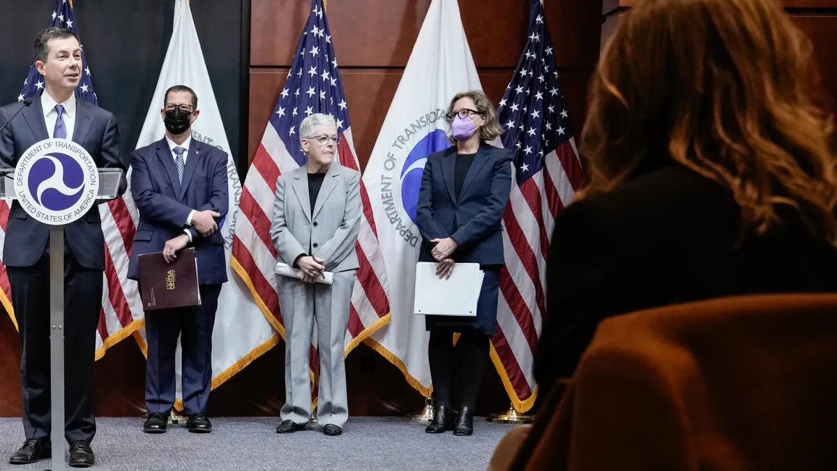 A group of people in business attire stand in front of the U.S. and  Department of Transportation flags as Secretary of Transportation Pete Buttigieg speaks behind a podium.