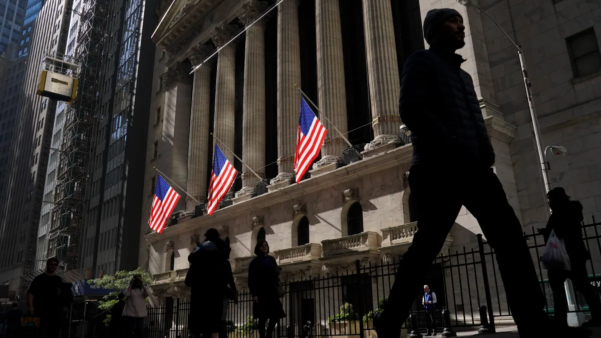 A sunlit New York Stock Exchanges is seen with 6 columns and 3 American flags with people walking by in shadow.