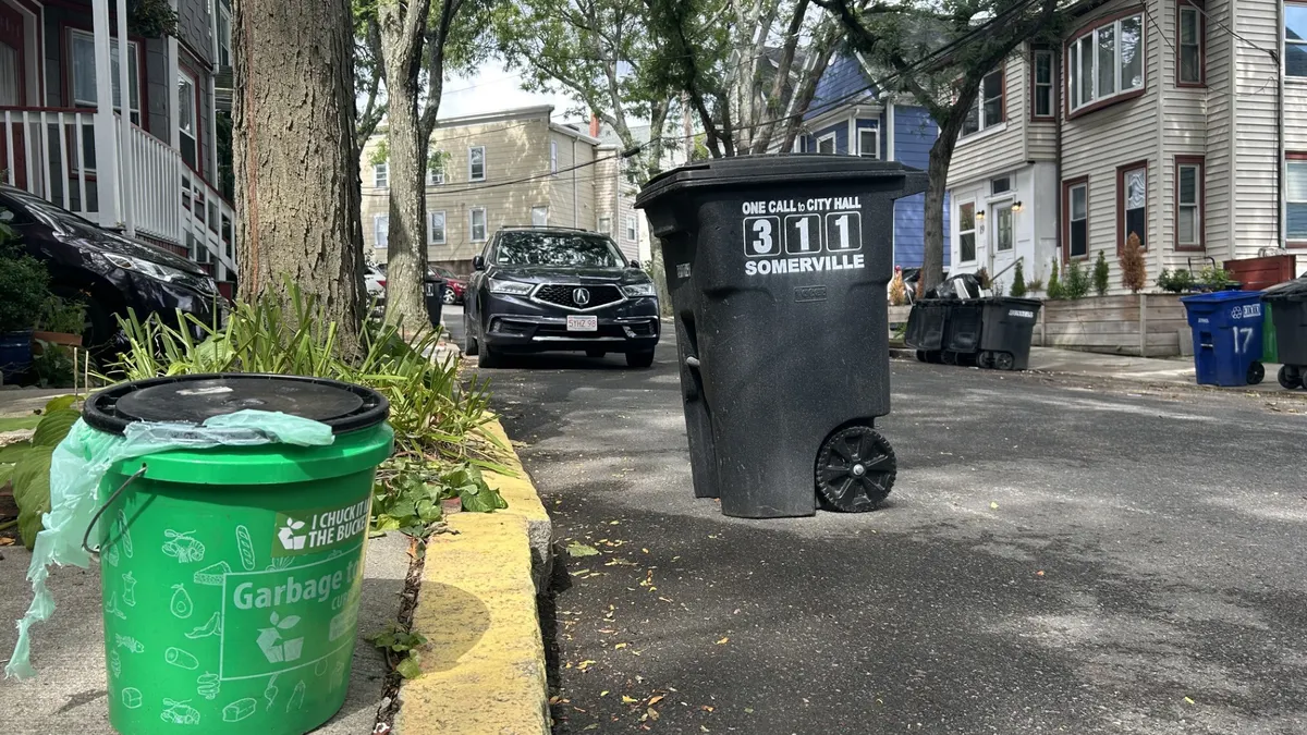 A small green bucket that says "Garbage to Garden" sits in the forefront of a street, with a large black bin and cars in the background.