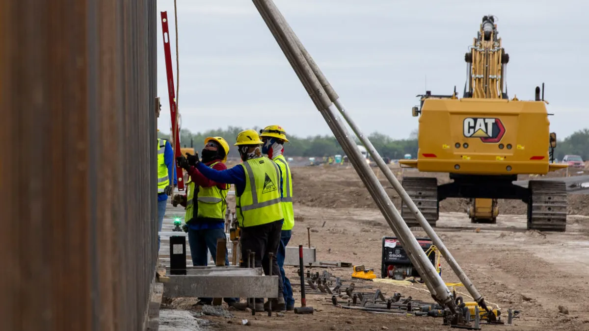 Three construction workers in bright helmets and vests stand next to a tall wall, with construction equipment in the background.