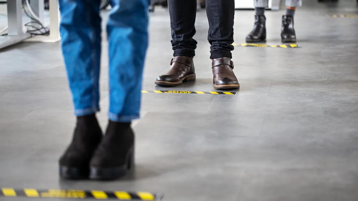 Low angle shot of business people standing behind social distancing signage on office floor. People in queue at office for safe entry checks post corona virus pandemic.