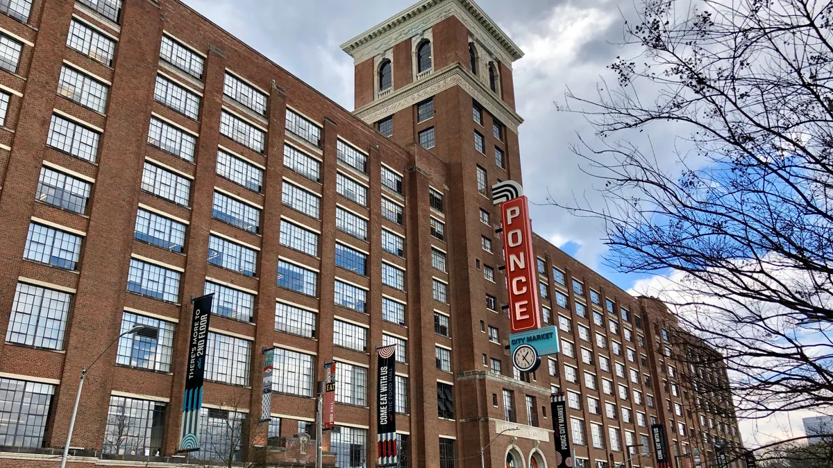 The many windows of a red brick building, seen at an angle, reflect the blue and clouds of the sky. A vertical red sign at the center reads "Ponce."