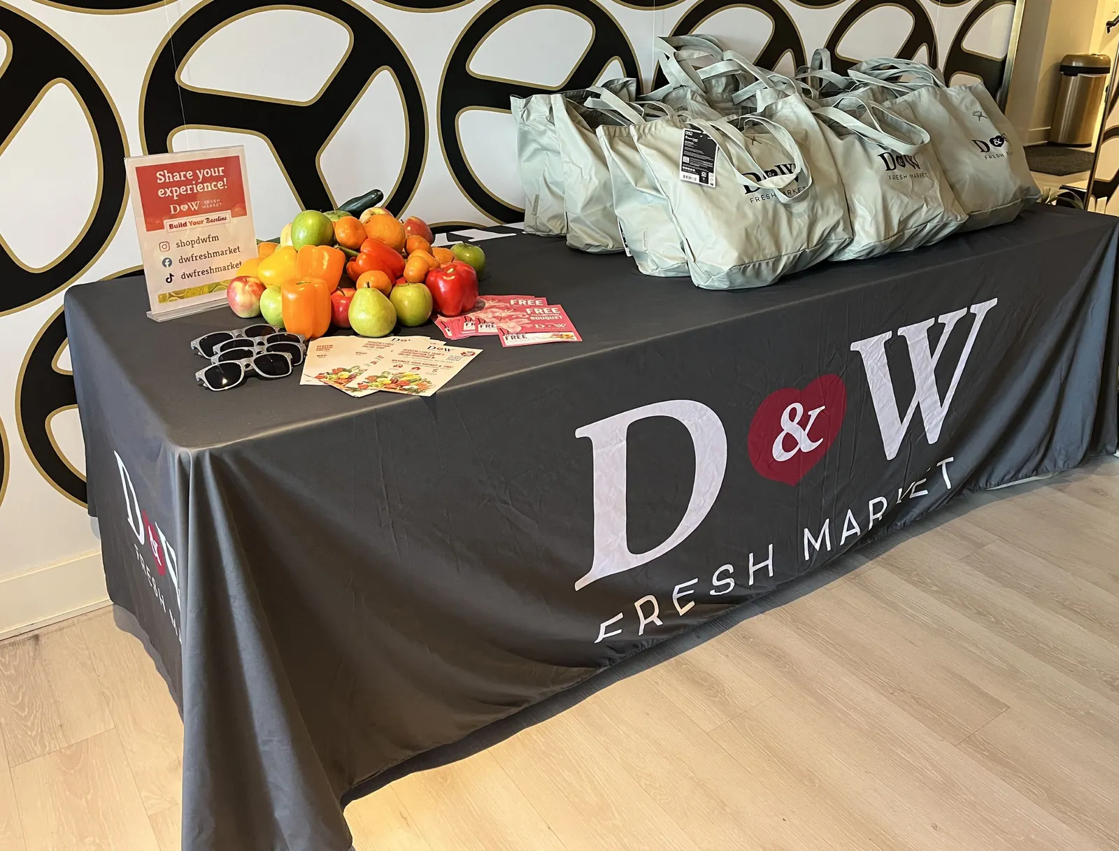 A table displaying fruit and vegetables to the left of a group of tote bags on a covering that reads "D&amp;W Fresh Market."