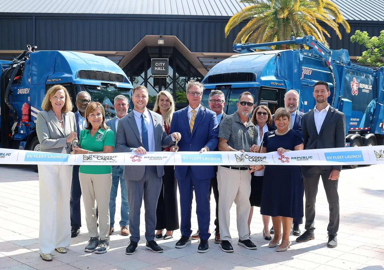 Several people wearing formal clothing stand at a ribbon cutting with Republic Services and Coconut Creek branding. Two waste collection trucks are parked behind them.