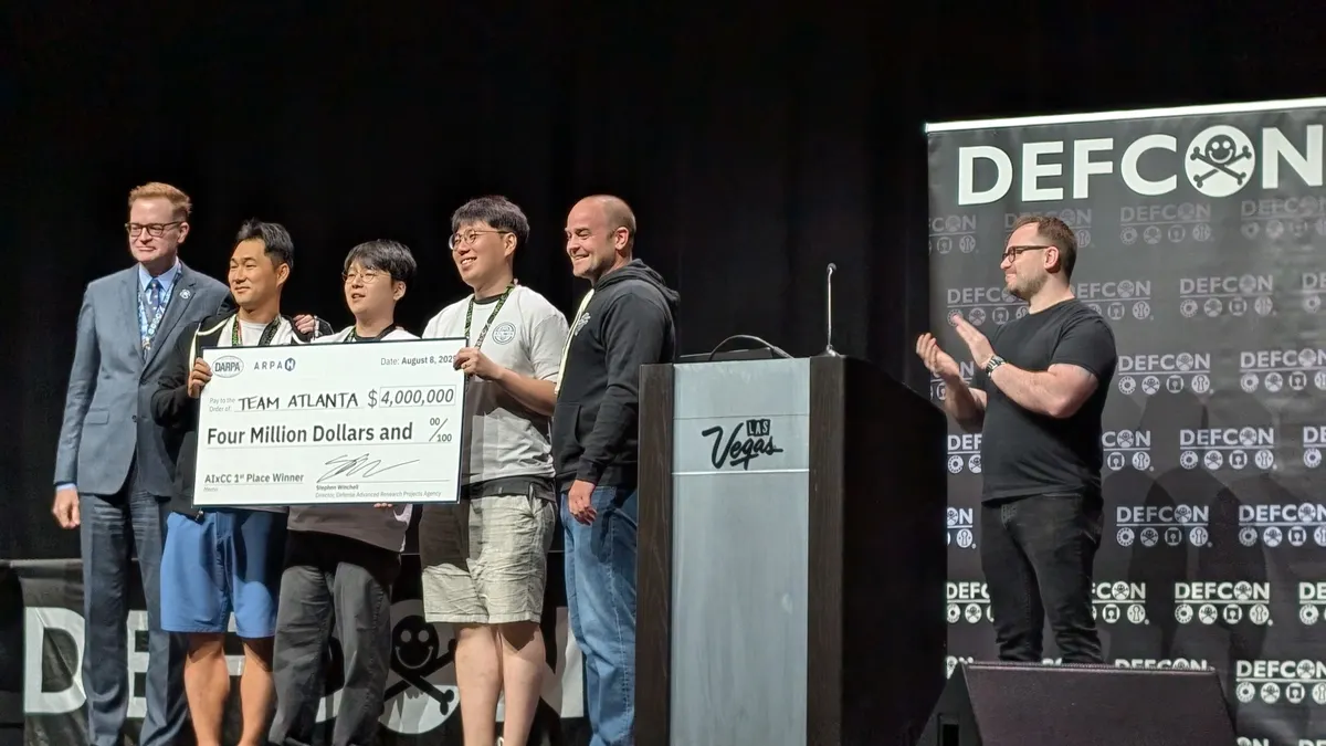 Six men stand on a stage. Three men hold an oversized $4 million check made out to "Team Atlanta," while two of the other men flank this trio and pose for a photo with them. The sixth person stands off to the side and applauds.