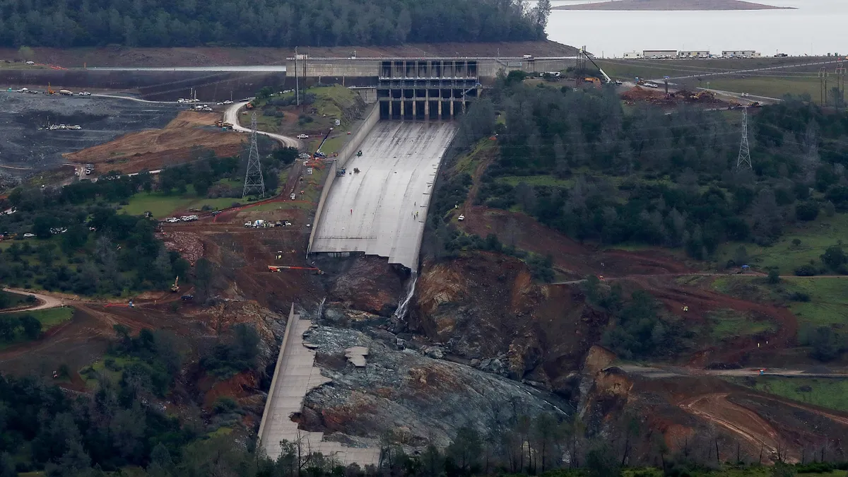 A dam sits amid mountain terrain with a lake in the background. The dam has a massive concrete chute extending from it that has extensive breakage.