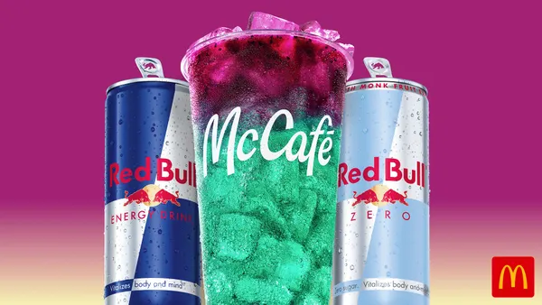 A close up photograph of a plastic cup with a green-blue liquid under pink liquid. The cup sits between two aluminum cans with Red Bull branding.
