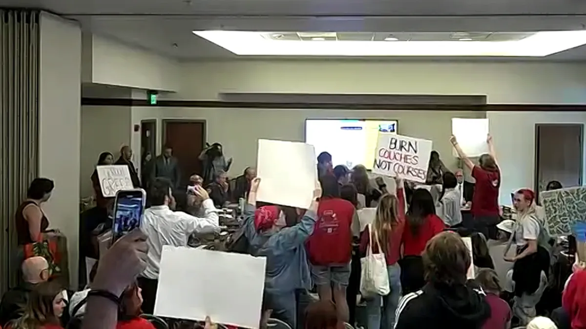 A board room is filled with young protestors holding signs and shouting.