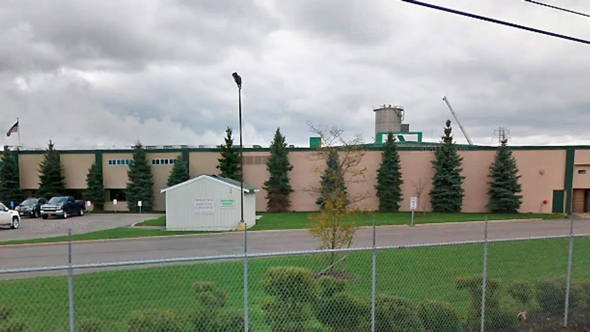 The exterior of an industrial facility with cars in a parking lot, which is surrounded by a chain-link fence.