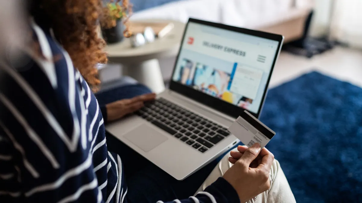 A person holds a credit card and sits with a laptop on her lap. The screen is open to show a shopping website.