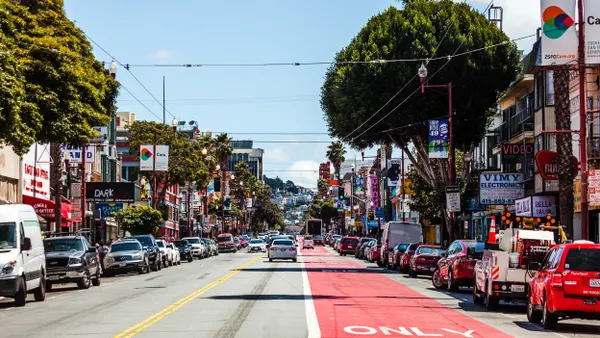 View down a two-lane street with a red-painted bike line on the right and parked cars on both side of the street, which is lined with small shops.