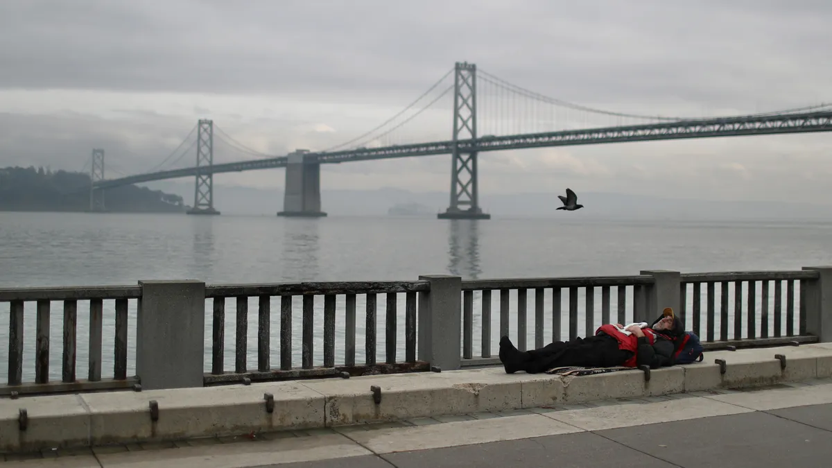 A person lies on the ground in front of a bridge over water.