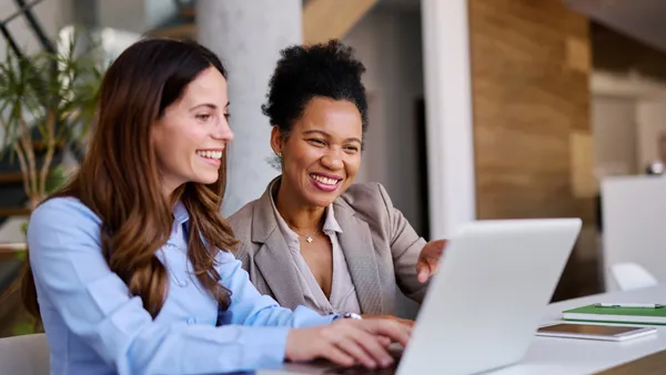Two businesswomen are collaborating on a project, looking at a laptop and smiling