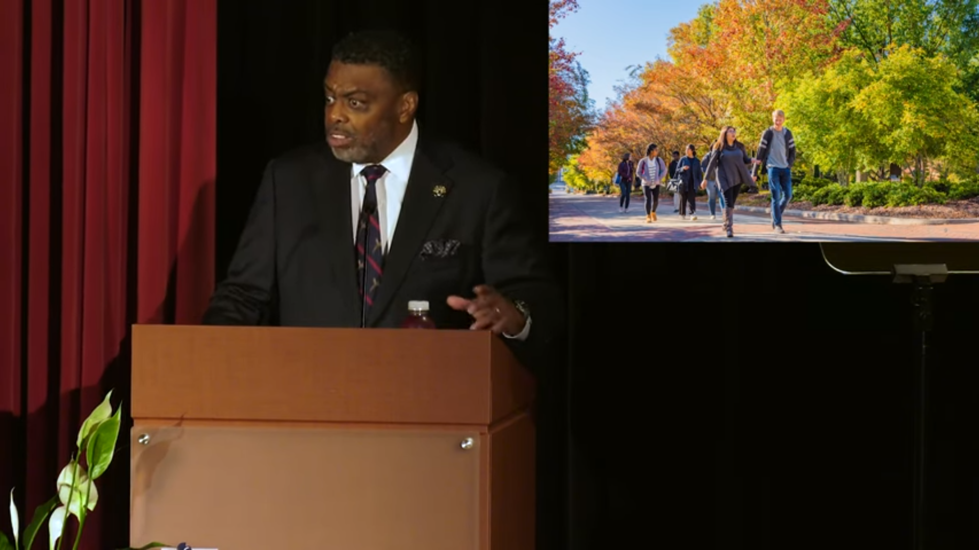 UNCG Chancellor Frank Gilliam speaks at a podium with a shot of students walking behind him.