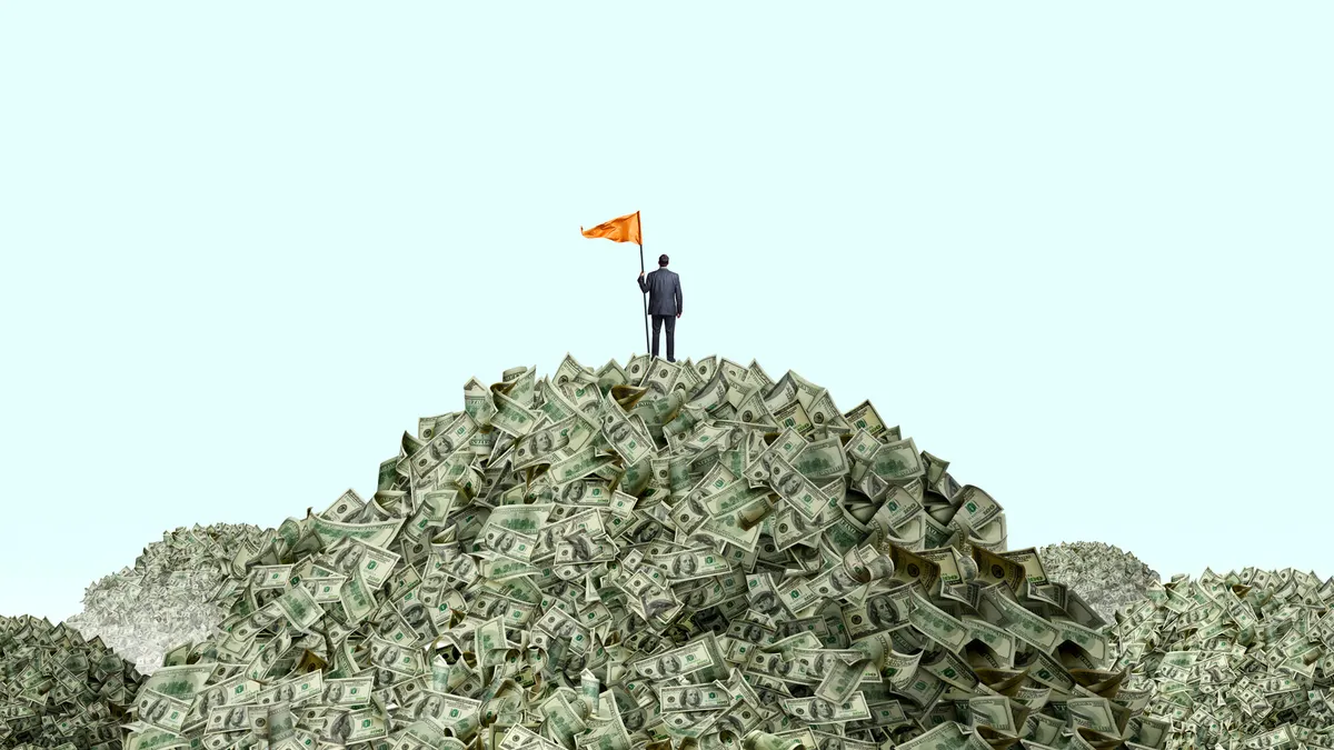 A businessman stands at the top of a mountain of money as he holds a large orange flag attached to a pole.