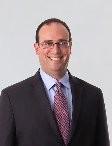 A smiling man in a dark suit, glasses and red patterned tie.