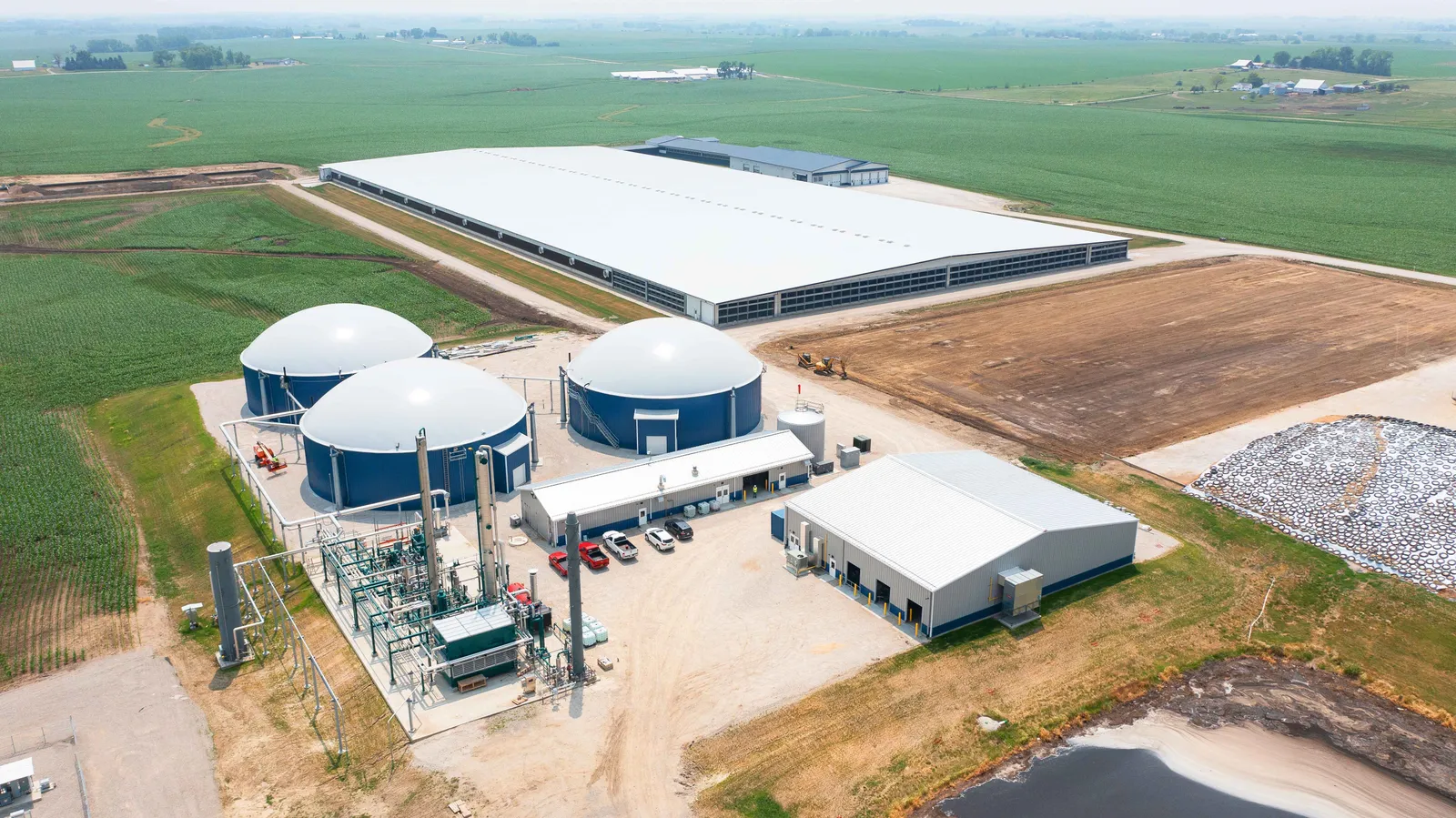 An industrial facility with three circular tank buildings sits near a vast shed and surrounding fields.