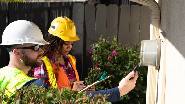 Two people look at an electric meter on the side of a house.