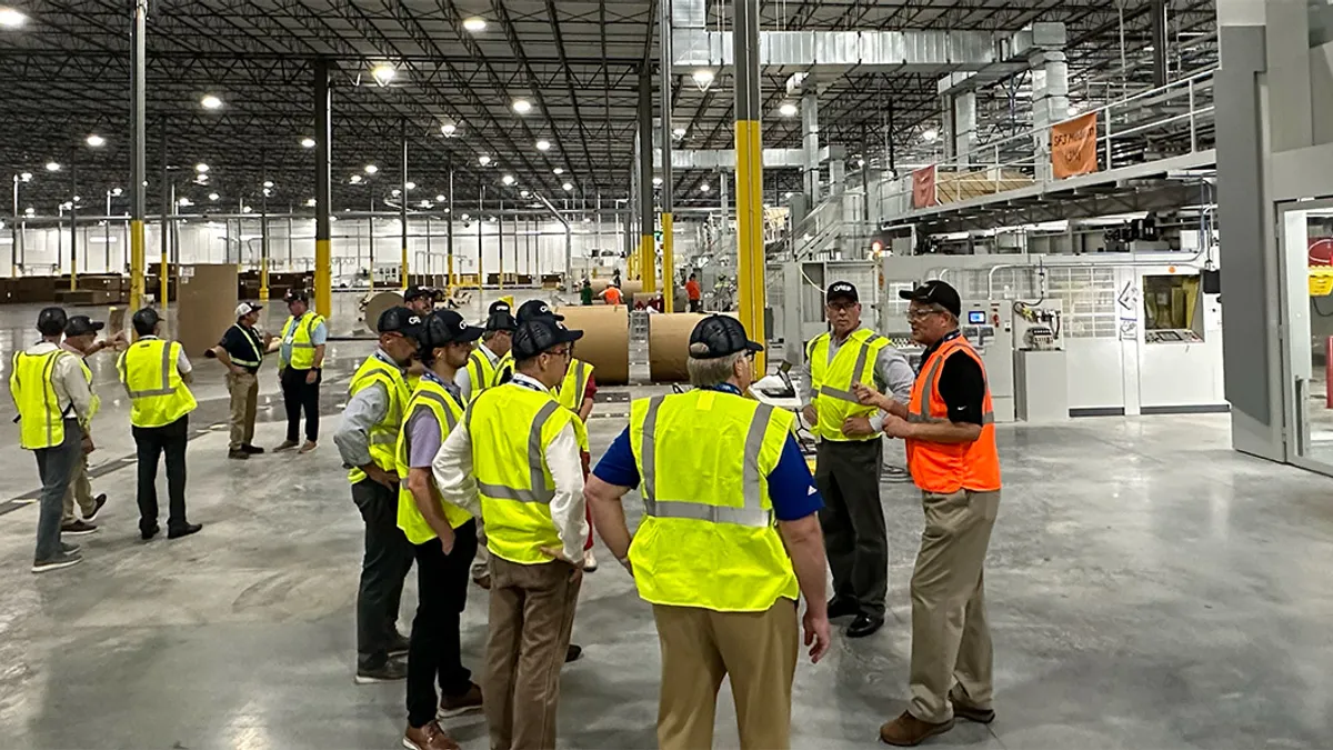 Fourteen people wearing safety vests have a discussion on the floor of an industrial facility.