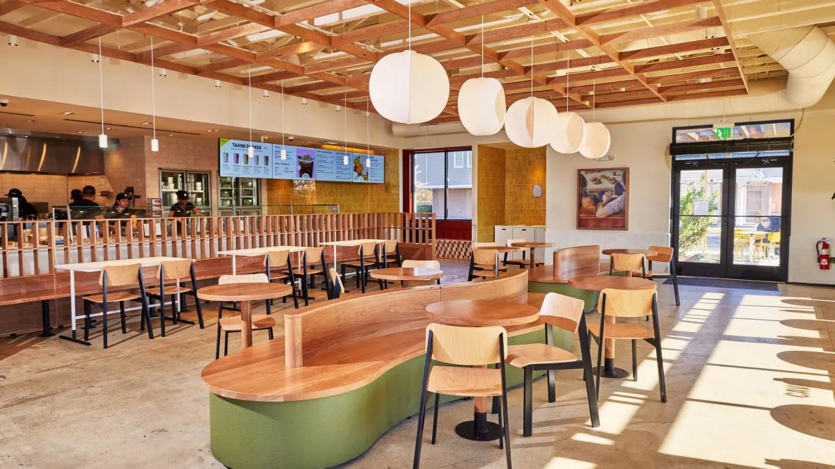 Wooden seats and tables inside a Cava. In the background there is a make line with digital menu boards.