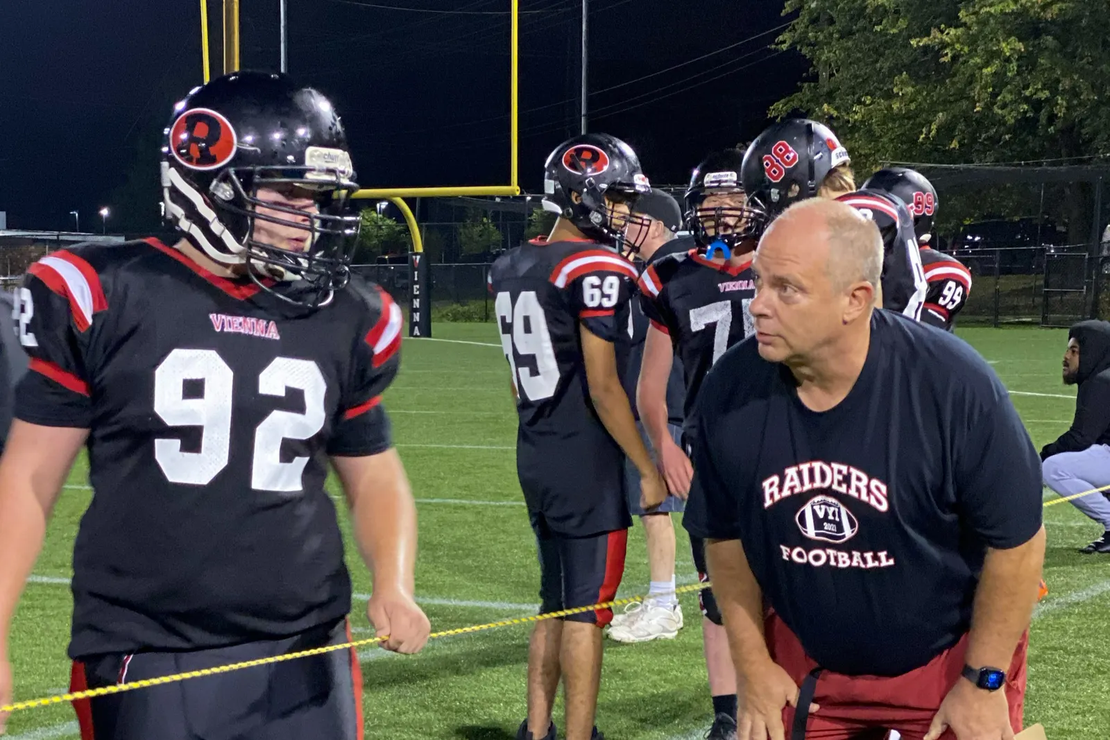 An adult is standing talkign with a football player near a football field at night.
